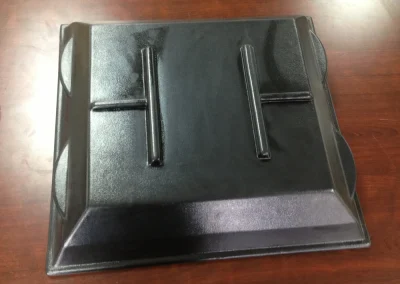 A black thermoformed plastic panel or cover with two raised T-shaped ribs and contoured edges, sitting on a wood table.