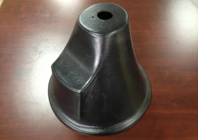 A black molded plastic bell-shaped cover or base with a small hole at the top and a contoured ridge, sitting on a dark wood table.