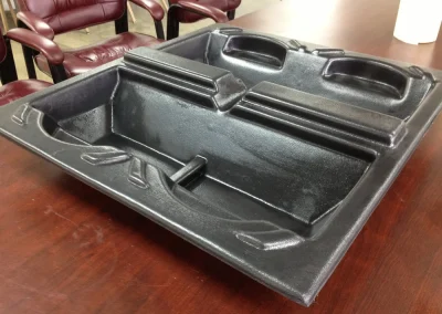 A black thermoformed plastic tray with molded compartments resting on a wood conference table, with maroon leather chairs in the background.