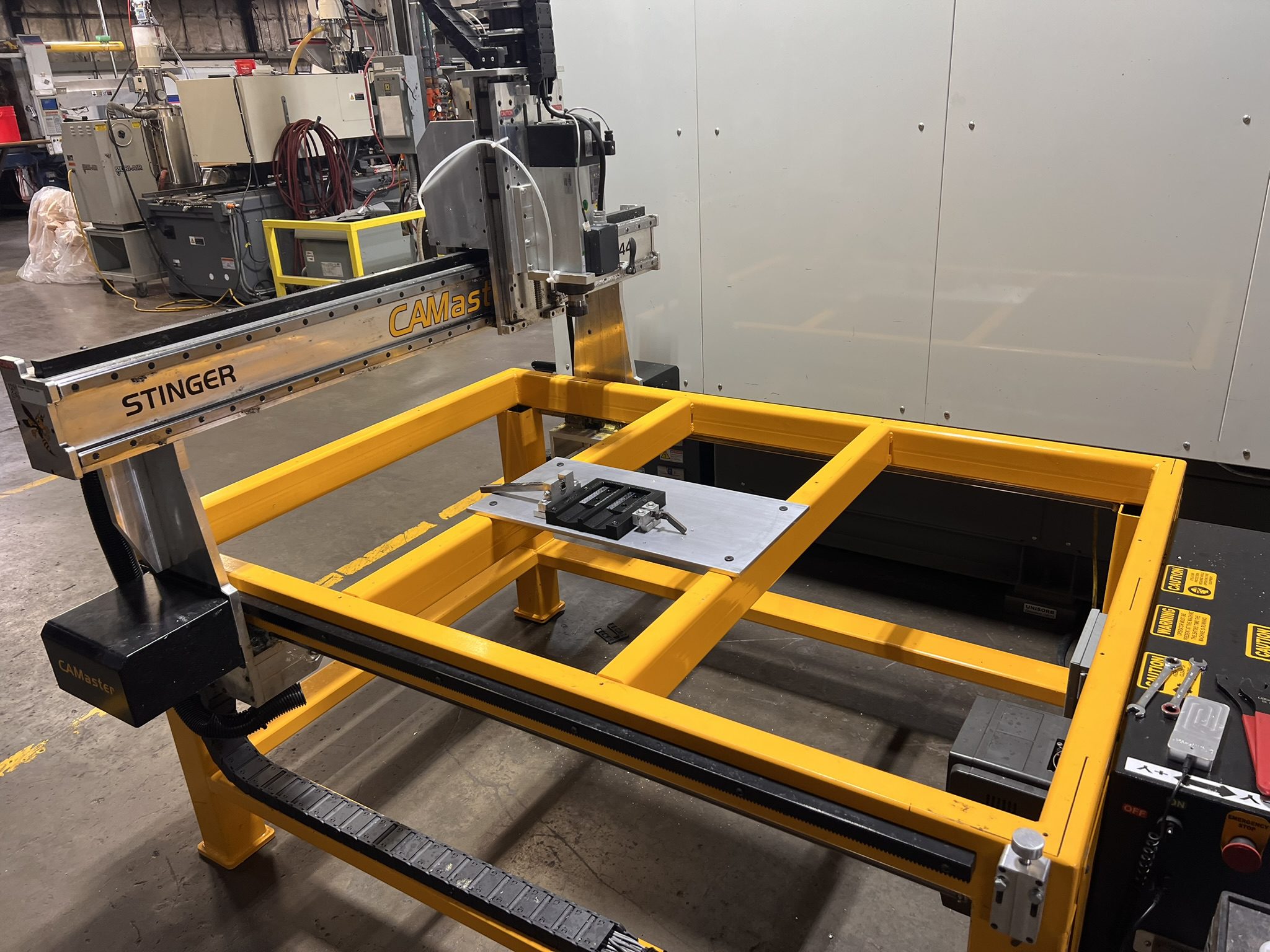 A CAMaster Stinger CNC router with a yellow steel frame and a fixturing plate on the bed, positioned on a manufacturing floor.