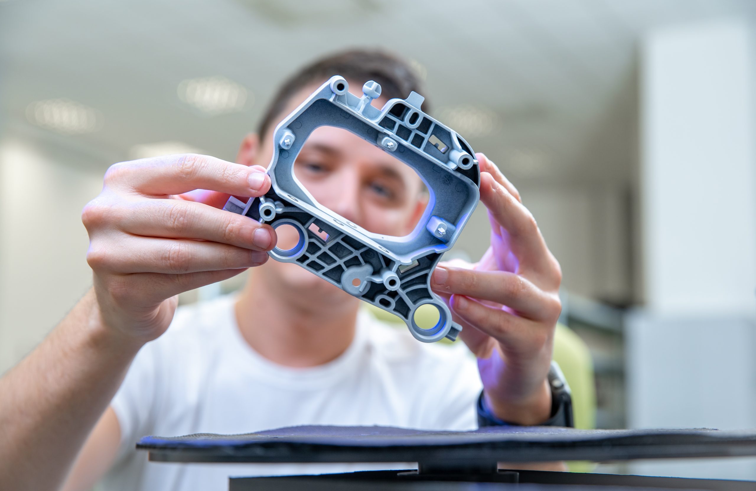 A person inspecting a complex injection-molded plastic and metal component, holding it up to examine it closely in a well-lit workspace.